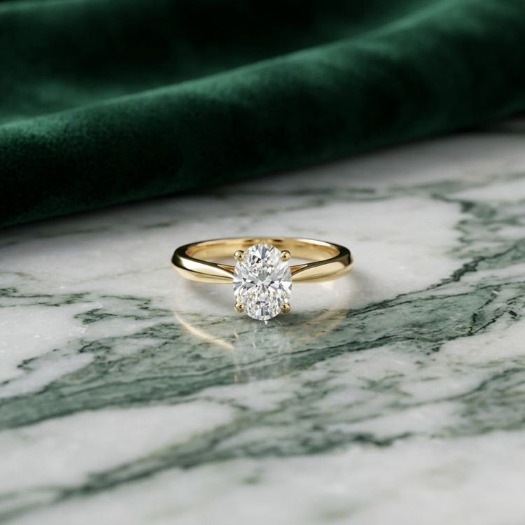 A 1/4 CT Diamond Oval Solitaire Ring in 14K gold is displayed on a white and green marble surface, with a green velvet cloth in the background.