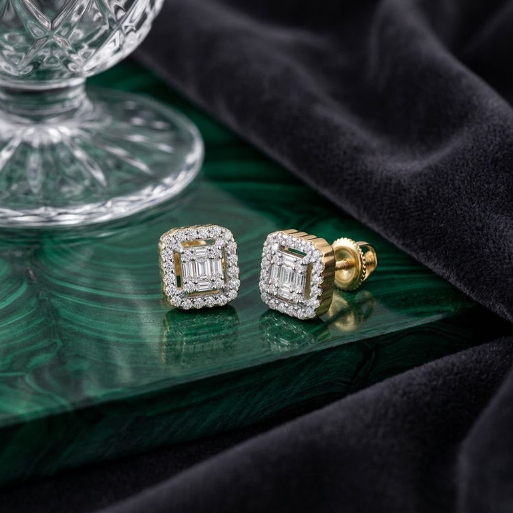 1/5 CTW Natural Diamond Baguette Earrings with a gold square double halo design rest on a green marbled surface beside a glass ornament and draped black velvet fabric.