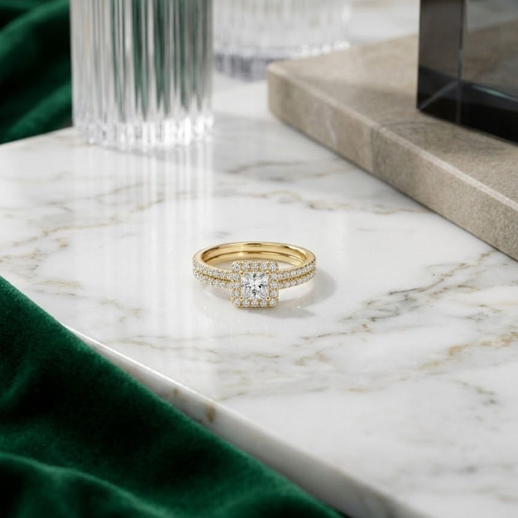 The 14k Gold Princess Diamond Square Halo Wedding Ring Set steals the spotlight, showcasing a striking square-cut princess diamond surrounded by a glittering halo and set on a band adorned with shimmering diamonds. Displayed gracefully on a white marble surface, the ring is complemented by a green velvet cloth, a glass, and a nearby stone coaster.