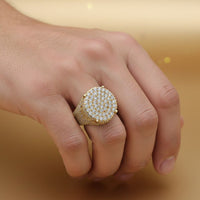A hand displays the Round Pavé Diamond Ring – 4.58 CT Gold Design, a large men’s gold ring adorned with sparkling white stones, worn on the ring finger against a soft golden background.