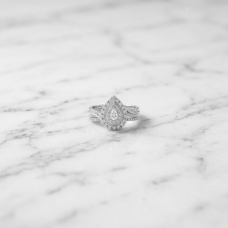 The 1/2 CTW Natural Diamond Pear Double Halo Bridal Set, featuring a teardrop-shaped diamond centerpiece surrounded by a double halo of smaller diamonds and set in silver, is elegantly displayed on a white marble surface.