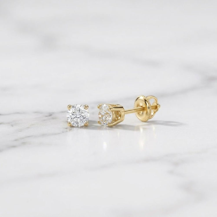 A pair of 14k Gold Round Diamond Solitaire Stud Earrings 1/5 CTTW with clear, round diamonds is displayed on a white marble surface.
