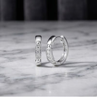 Two 10k gold diamond fashion hoop earrings (1/10CTW), featuring rectangular, diamond-studded fronts, are displayed on a polished white marble surface with a softly blurred dark background. These elegant natural diamond earrings catch the light beautifully, making them a timeless addition to any ensemble.