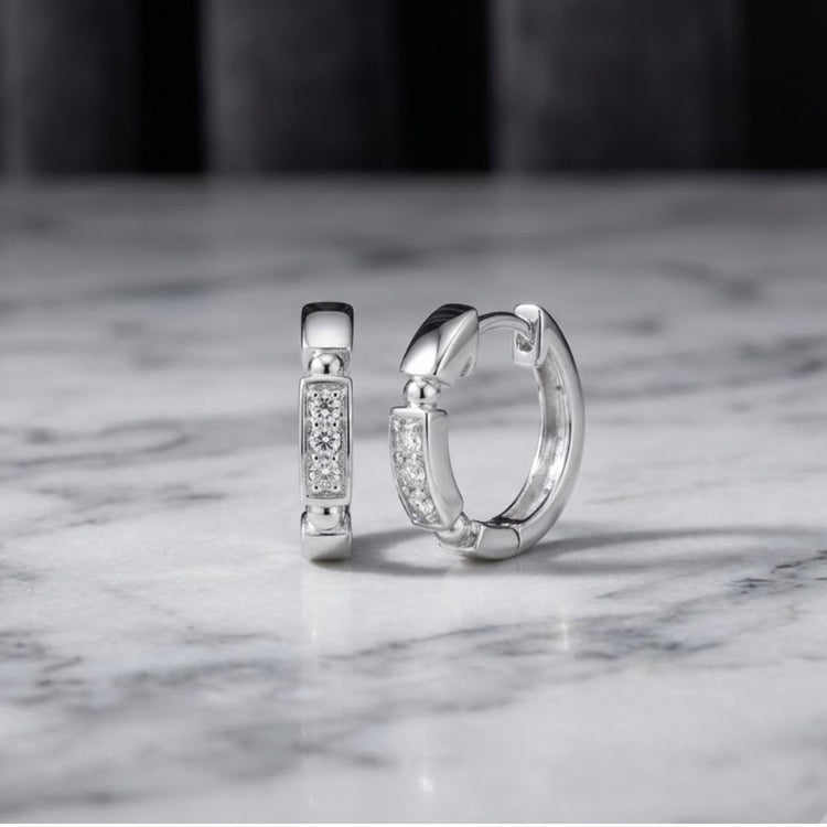 Two 10k gold diamond fashion hoop earrings (1/10CTW), featuring rectangular, diamond-studded fronts, are displayed on a polished white marble surface with a softly blurred dark background. These elegant natural diamond earrings catch the light beautifully, making them a timeless addition to any ensemble.