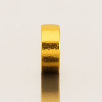 The 8mm Flat Band Ring - 24k is a plain, thick gold band standing upright on a reflective light surface, its smooth yet subtly textured finish casting a soft shadow below.