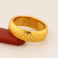 A close-up of the 8mm Hammered Band Ring - 24k, featuring a textured hammered surface, is shown resting against smooth curved red material on a light background.