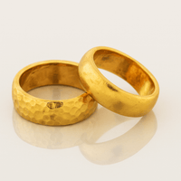 An 8mm Hammered Band Ring in 24k gold with a textured finish rests beside a smooth, comfort-fit 24k gold wedding band on a light surface.