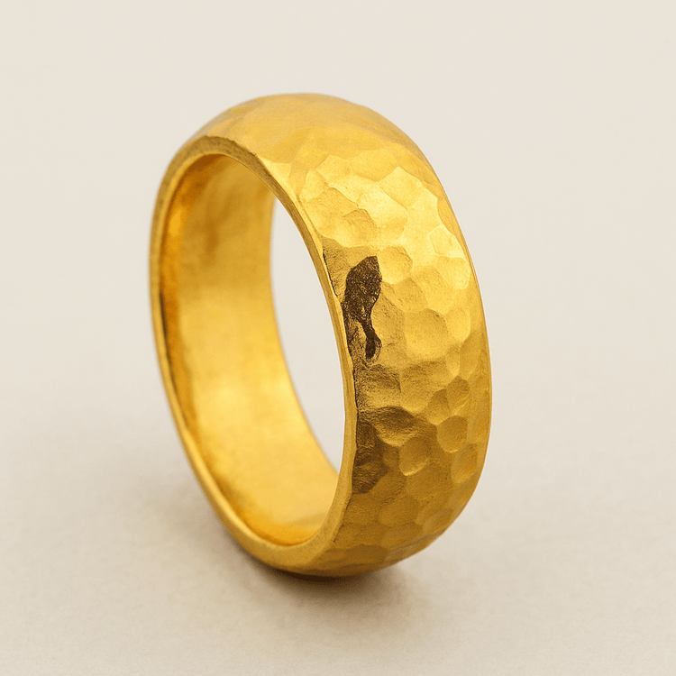 The 8mm Hammered Band Ring - 24k features a wide, textured matte gold design with a comfort fit and a unique dark accent, shown standing upright on a neutral background.