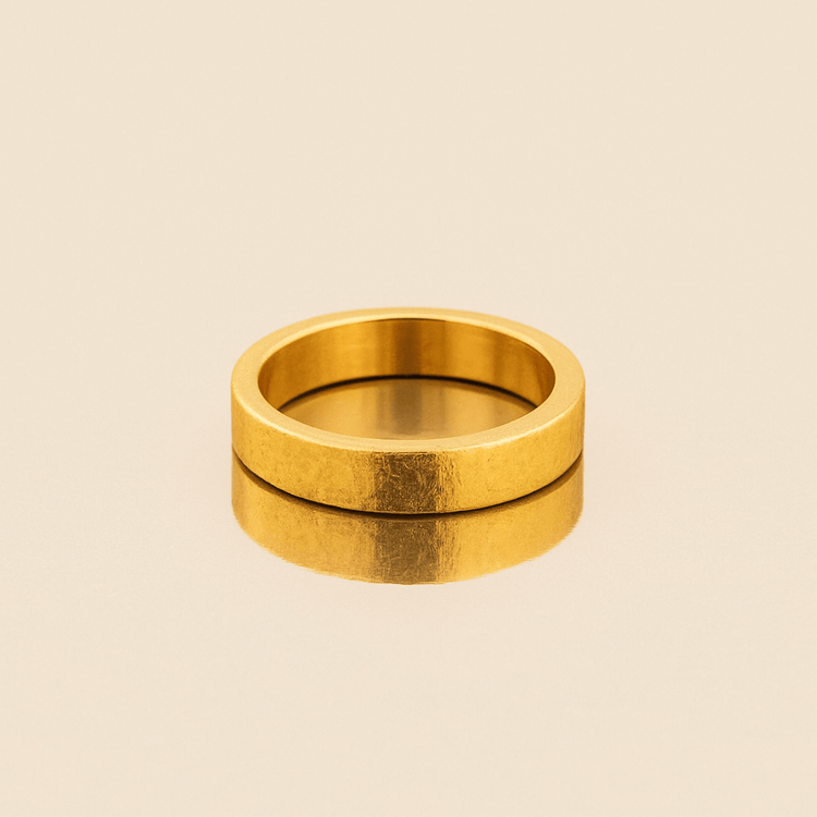 The 4mm Flat Band Ring - 24k, a sleek and polished gold wedding band with flat edges, rests on a reflective light beige surface, casting a soft reflection below.