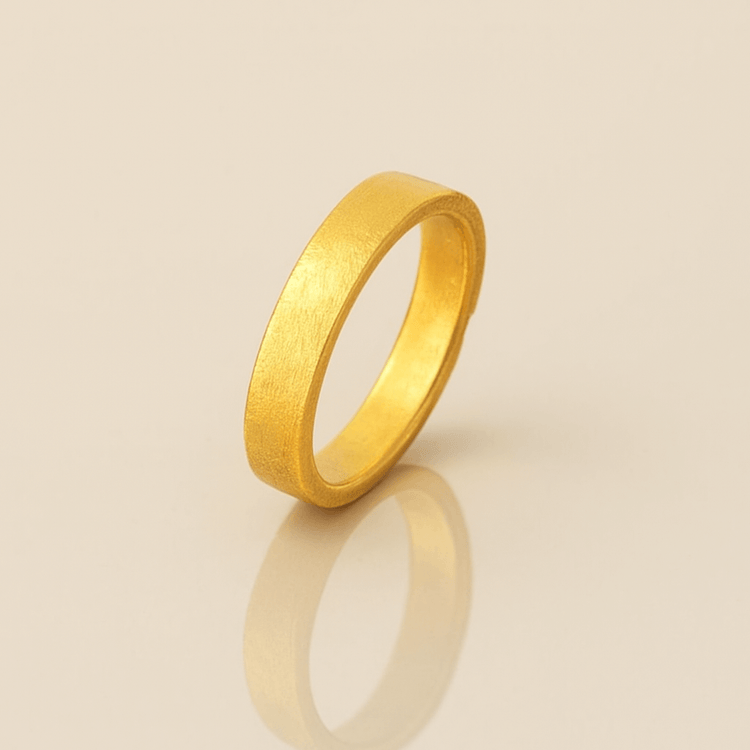 The 4mm Flat Band Ring - 24k features a simple matte finish and stands upright on a shiny, light surface, casting a soft reflection below.