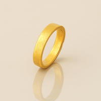 The 4mm Flat Band Ring - 24k features a simple matte finish and stands upright on a shiny, light surface, casting a soft reflection below.