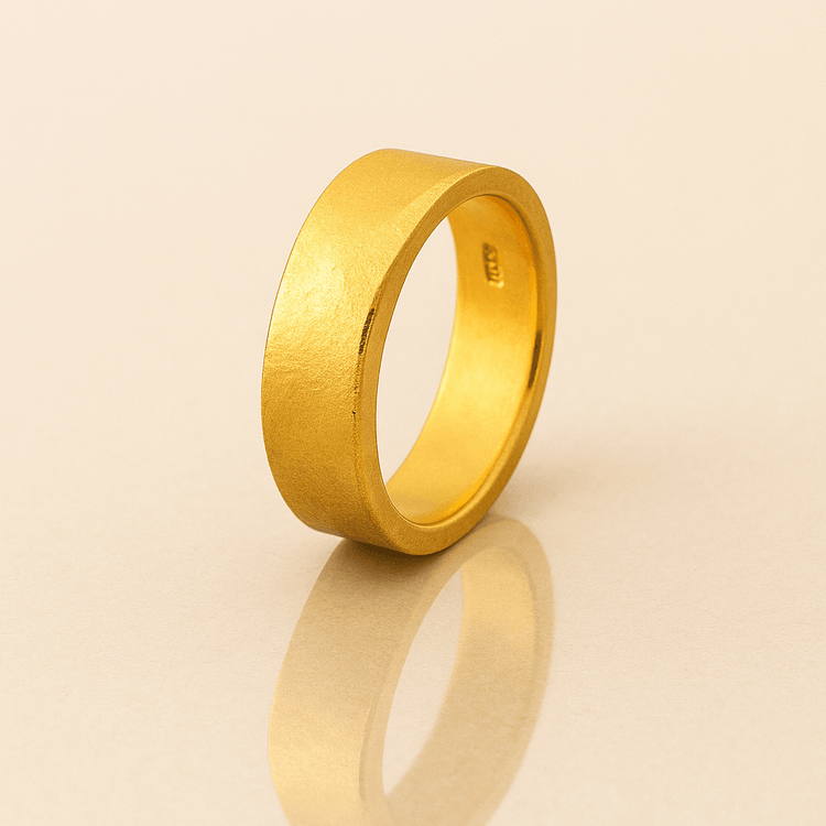 The 6mm Flat Band Ring -24k features a simple, wide matte gold finish and stands upright on a light, reflective surface, casting a soft shadow and reflection.