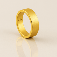 The 6mm Flat Band Ring -24k features a matte finish and "24K" engraving on the inner band, displayed upright on a reflective beige surface.