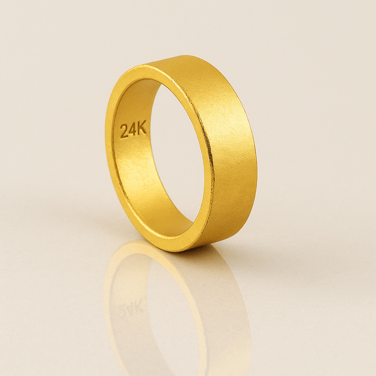 The 6mm Flat Band Ring -24k features a matte finish and "24K" engraving on the inner band, displayed upright on a reflective beige surface.