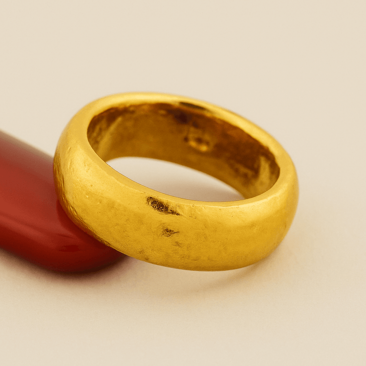 The 8mm Band Ring -24k, featuring a smooth, gently worn surface and classic comfort-fit design, rests against a red curved object on a neutral beige background, highlighting its timeless elegance.