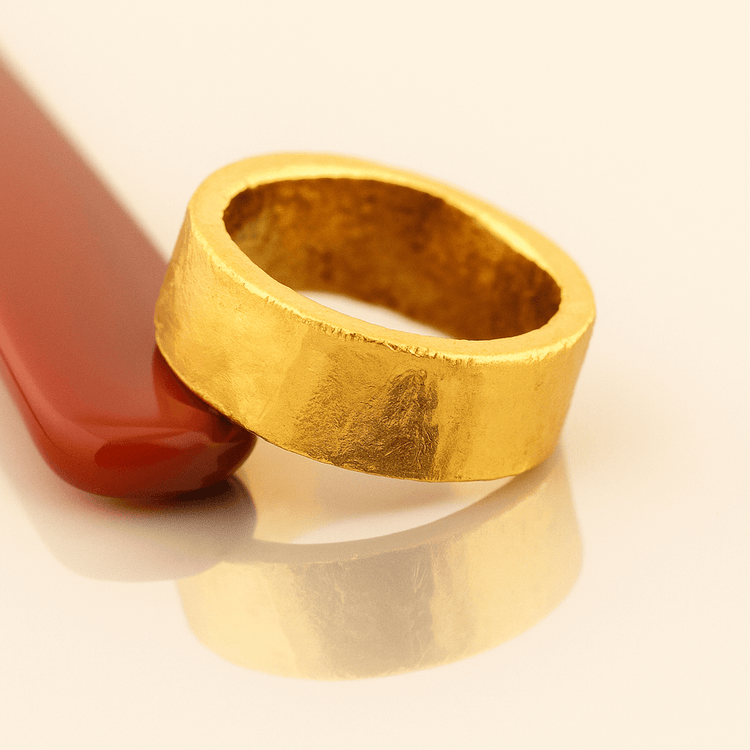 The 10mm 1oz Flat Band Ring-24k, with a chunky matte gold finish, rests against a smooth red object on a glossy cream-colored surface, casting a subtle reflection below.