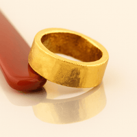 The 10mm 1oz Flat Band Ring-24k, with a chunky matte gold finish, rests against a smooth red object on a glossy cream-colored surface, casting a subtle reflection below.