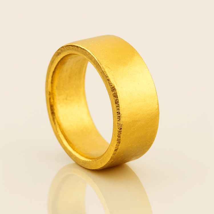 The 10mm 1oz Flat Band Ring-24k features a wide, flat matte gold band with a minimalist design, displayed upright on a reflective beige surface.