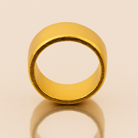 The 10mm 1oz Flat Band Ring-24k stands upright on a reflective surface with a light beige background, casting a subtle shadow and reflection that highlight its minimalist design.