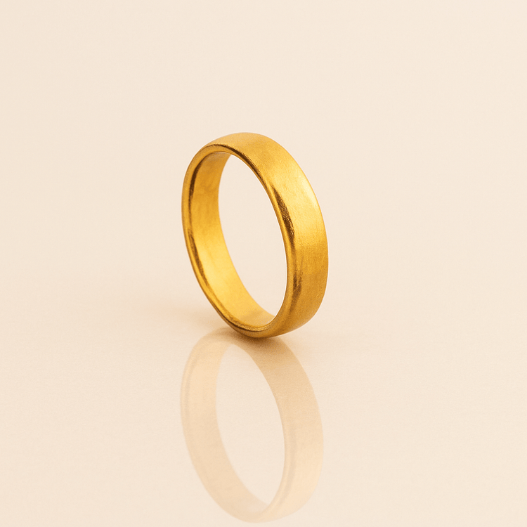 The 6mm Band Ring 24k, a sleek and polished gold band, stands upright on a light beige surface, casting a gentle reflection below.
