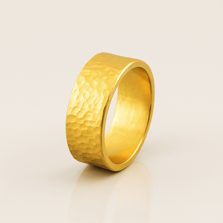 The 8mm, 24k Hammered Flat Band stands upright on a neutral, light-colored surface, highlighting the elegance of its hammered texture and flat design.