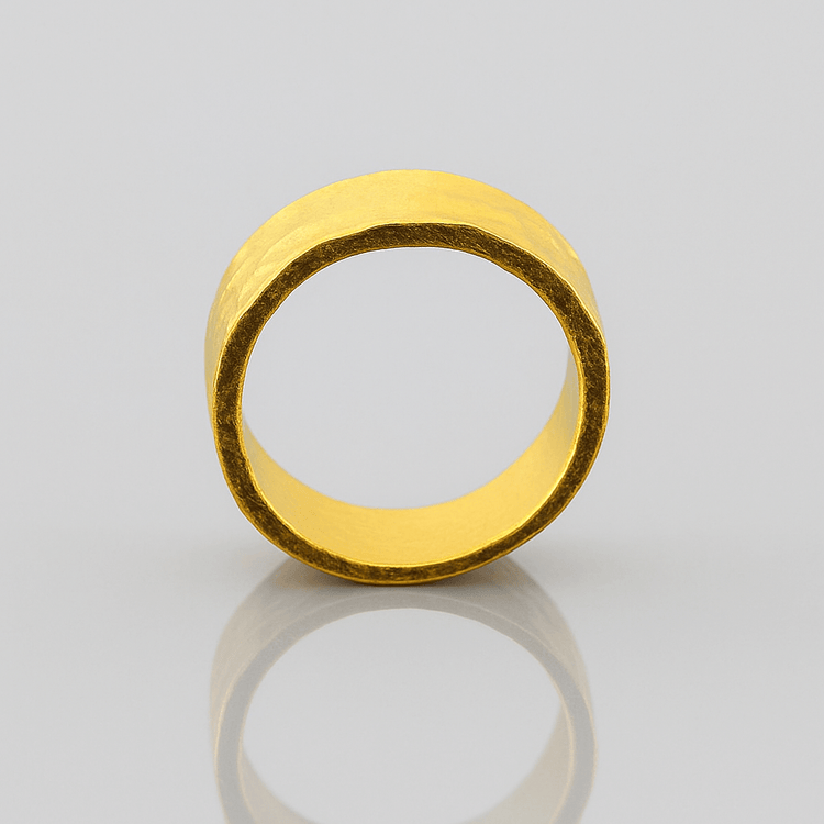 The 8mm, 24k Hammered Flat Band stands upright on a reflective light gray surface, showcasing its matte, uneven texture and rich gold color.
