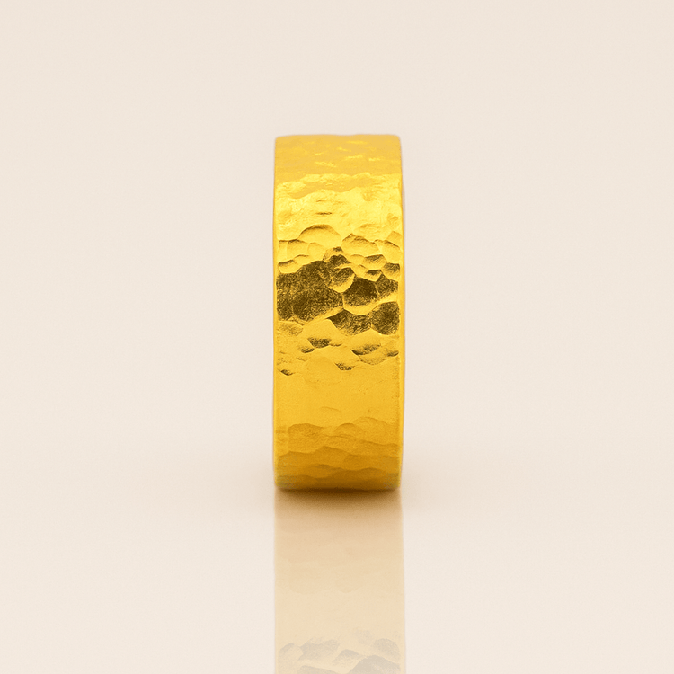 The 8mm, 24k Hammered Flat Band features a bold, wide profile and unique hammered texture, standing upright on a neutral, reflective surface to showcase its striking presence.