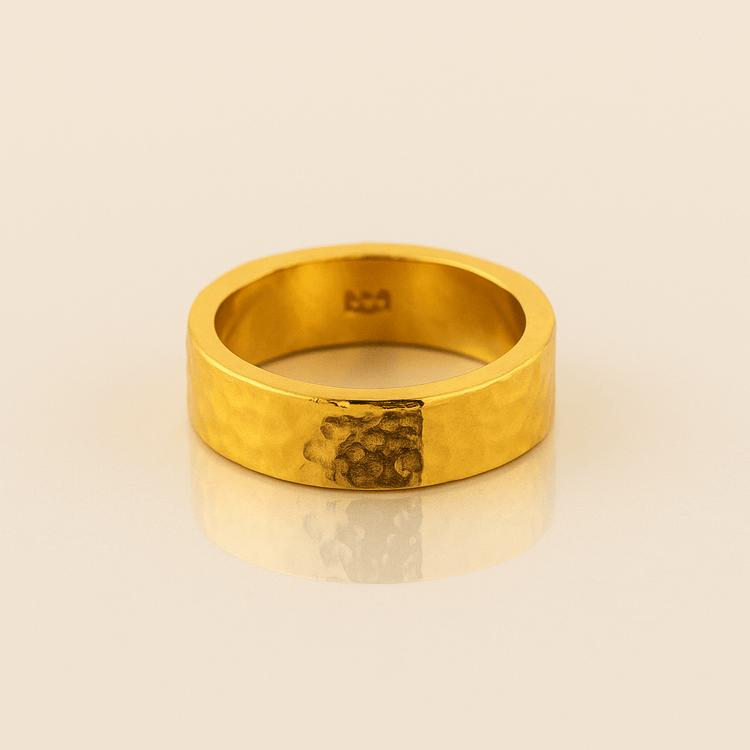 The 6mm, 24k Hammered Flat Band features a comfort-fit design with a unique darker hammered section on one side, displayed on a light beige surface.