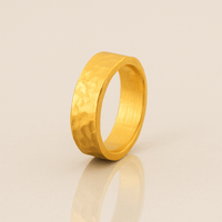 The 6mm, 24k Hammered Flat Band stands upright on a smooth, reflective beige surface, showcasing its comfort-fit design and wide, textured band.