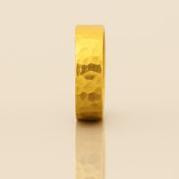 The 6mm, 24k Hammered Flat Band stands upright on a reflective beige surface, its gold finish casting a subtle shadow below.