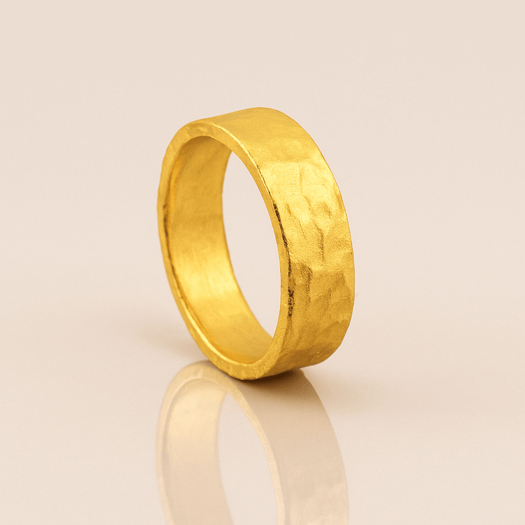 The 6mm, 24k Hammered Flat Band stands upright on a light beige reflective surface, casting a gentle shadow and reflection.
