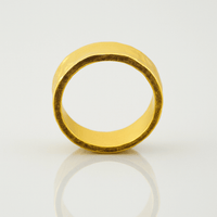 The 6mm, 24k Hammered Flat Band features a simple, wide gold design with a matte finish and stands upright on a reflective white surface, creating a subtle reflection beneath it.