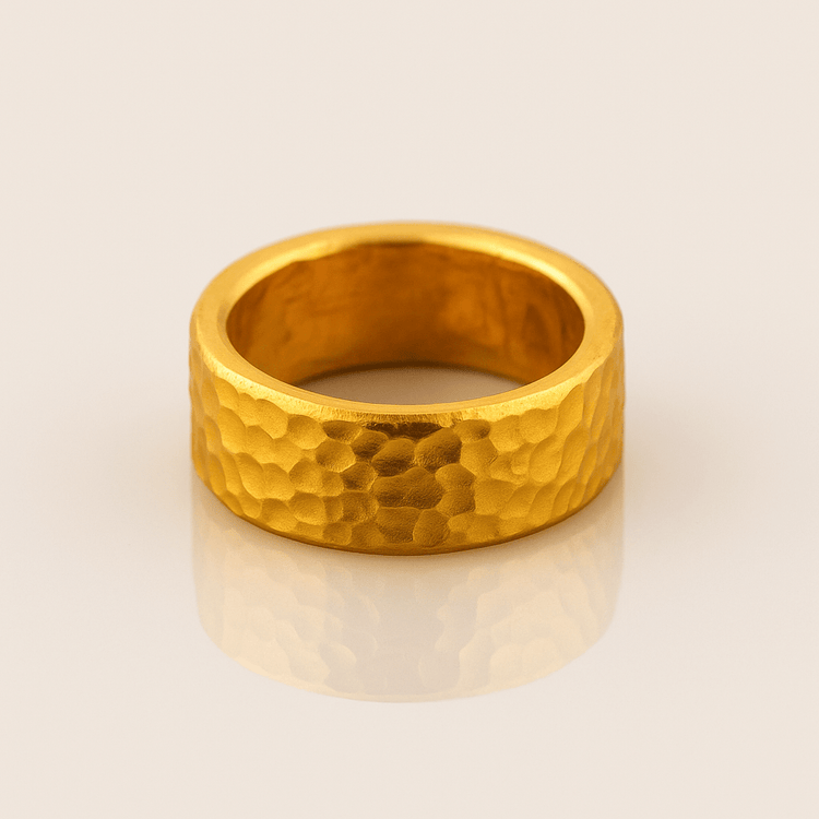 The 10mm, 24k 1oz Hammered Flat Band features a matte, faceted texture and wide profile, displayed on a reflective surface to highlight its warm metallic sheen and comfortable, striking comfort-fit design.