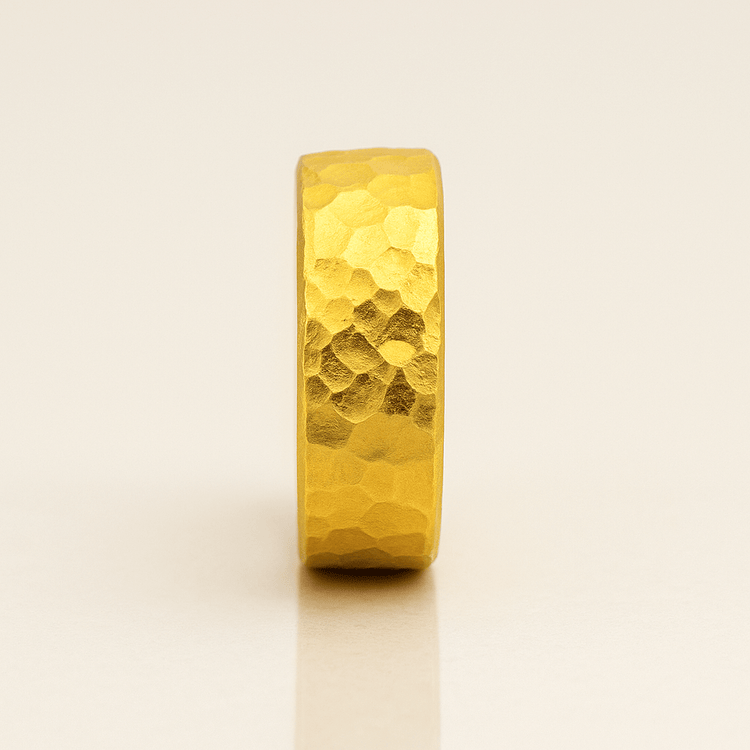 The 10mm, 24k 1oz Hammered Flat Band with a comfort-fit design stands upright on a light, reflective surface against a neutral background.