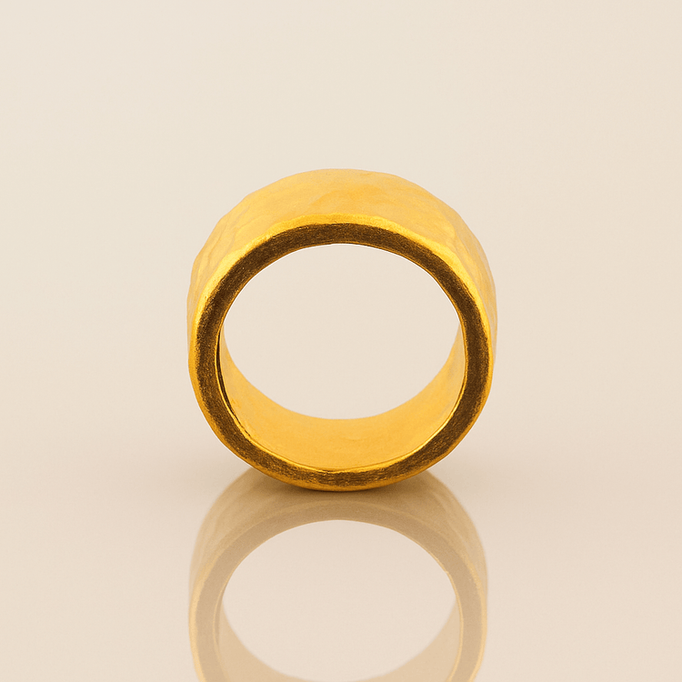 The 10mm, 24k 1oz Hammered Flat Band features a matte finish, smooth edges, and a comfort-fit design for everyday elegance. It stands upright on a neutral, reflective surface, showcasing its wide hammered texture.