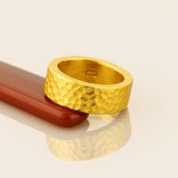 The 10mm, 24k 1oz Hammered Flat Band features a textured matte finish and artisanal design. Set on a glossy, reddish-brown cylinder against a beige background, the comfort-fit ring casts a soft shadow.