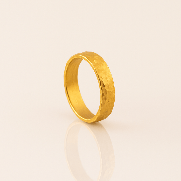 The 4mm, 24k Hammered Band Ring stands upright on a light, reflective surface, showing its unique texture and a subtle reflection, with an off-white background highlighting the comfort-fit design.