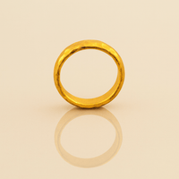 The 4mm, 24k Hammered Band Ring stands upright on a smooth beige surface, its comfort-fit design and textured gold finish clearly reflected beneath it.