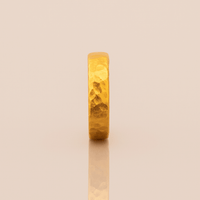 The 4mm, 24k Hammered Band Ring stands upright on a reflective, light beige surface, showcasing its comfort fit and elegant hammered texture.