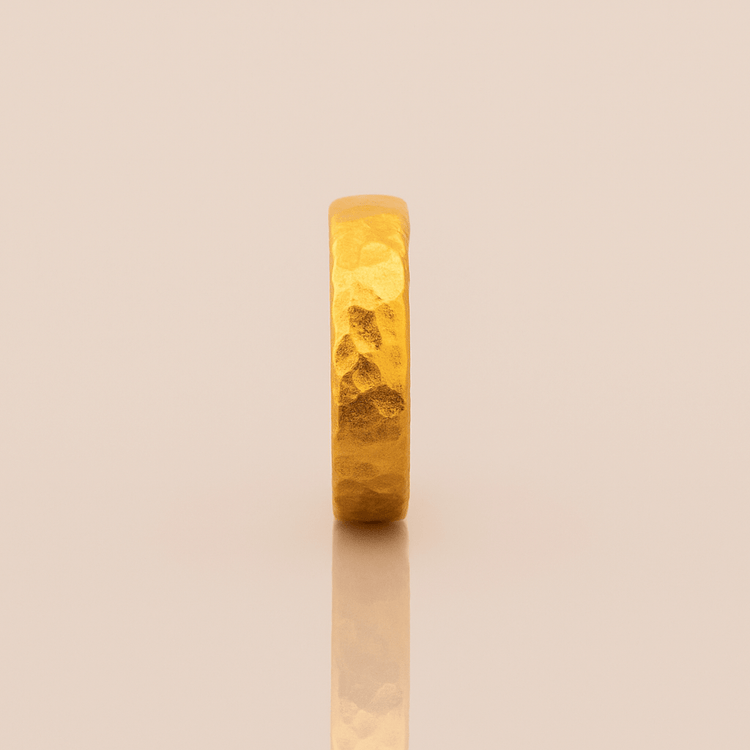 The 4mm, 24k Hammered Band Ring stands upright on a reflective, light beige surface, showcasing its comfort fit and elegant hammered texture.