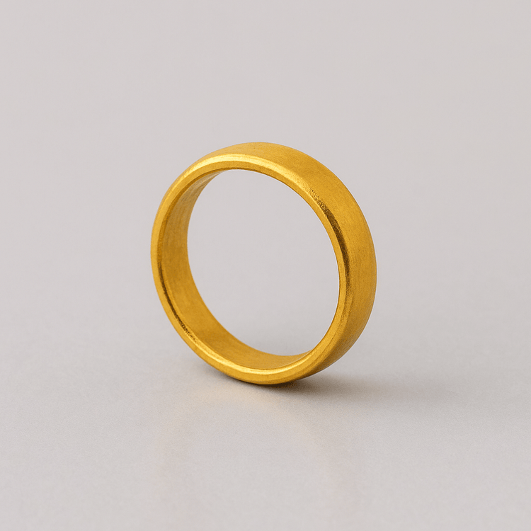 The 4mm, 24k Band Ring features a smooth, plain gold finish and stands upright on a light gray surface against a neutral background.