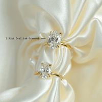 Two 2.12ct Oval Lab Diamond Engagement Rings in 14k yellow gold rest on ivory satin, their hidden halos and elegant settings enhancing the lab-grown diamonds' sparkle.