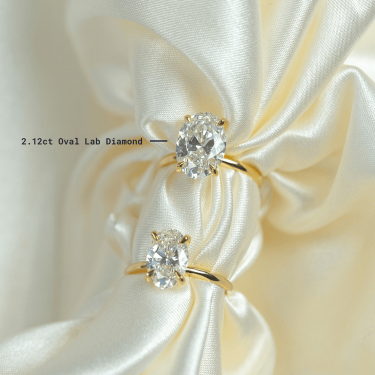 Two 2.12ct Oval Lab Diamond Engagement Rings in 14k yellow gold rest on ivory satin, their hidden halos and elegant settings enhancing the lab-grown diamonds' sparkle.