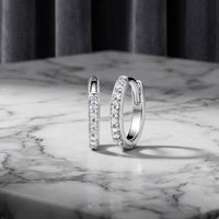 A pair of 1/10ctw Natural Diamond Hoop Earrings in 10K Gold is displayed on a white marble surface with gray veining, while a gray curtain hangs softly blurred in the background.