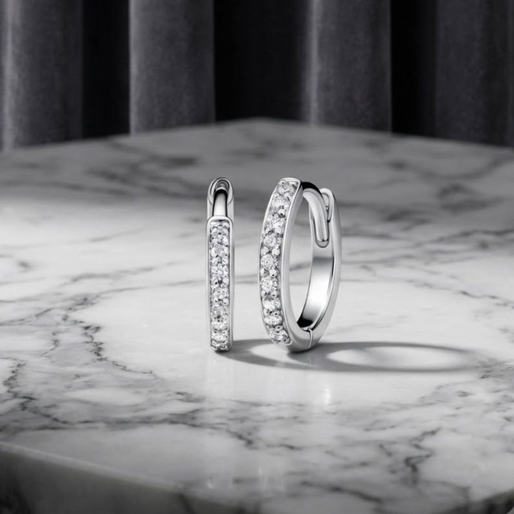 A pair of 1/10ctw Natural Diamond Hoop Earrings in 10K Gold is displayed on a white marble surface with gray veining, while a gray curtain hangs softly blurred in the background.