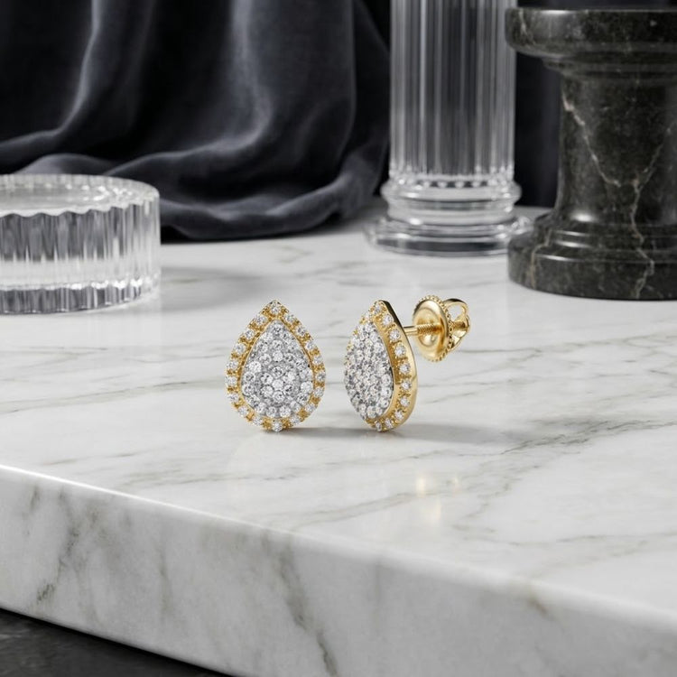 A pair of 1/4 CTW Natural Diamond Pear Earrings in 10K gold are displayed on a white marble surface, with decorative glass and stone objects in the background.