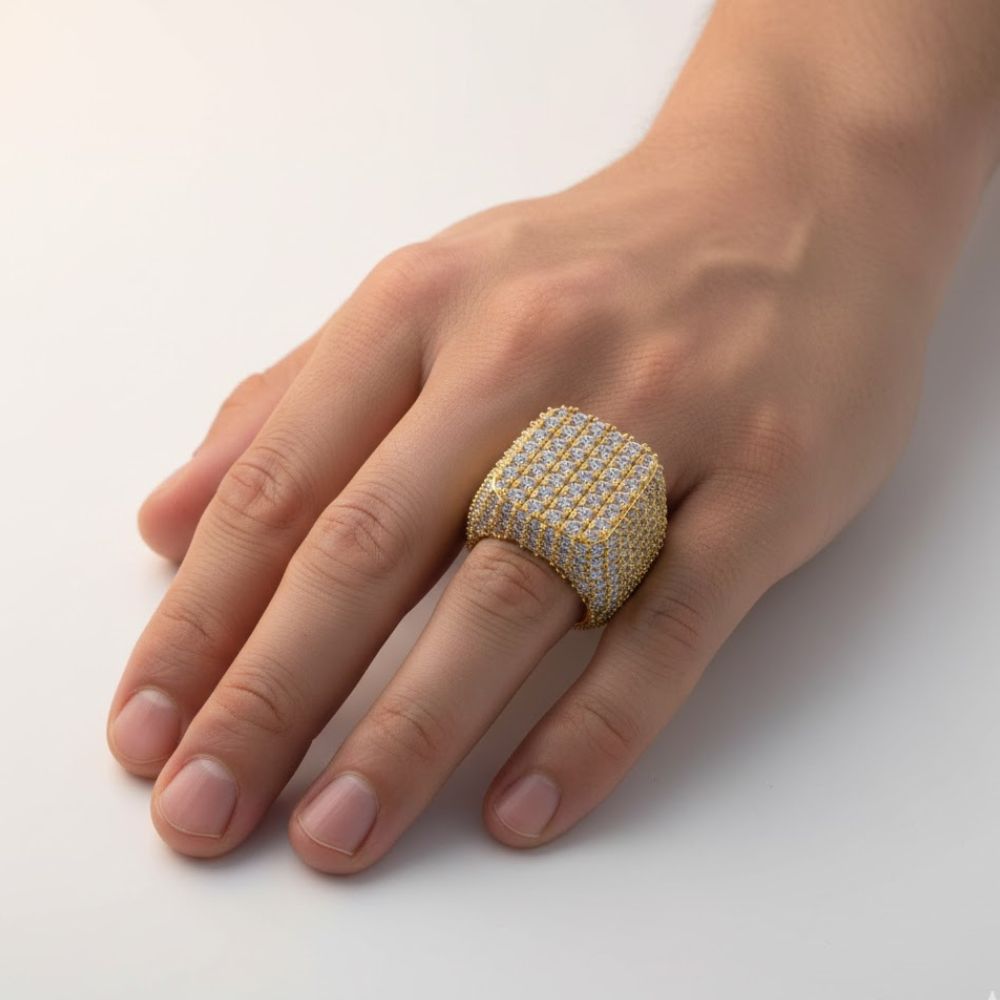 A hand showcases the Square Pavé Diamond Ring – 18 CT Gold Statement, a bold, ornate men’s ring encrusted with diamonds. Its striking, square design covers most of the finger and stands out against a plain background.