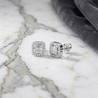 A pair of 1/5 CTW Natural Diamond Baguette Earrings with halo settings rests on a white marble surface beside a folded piece of dark gray velvet fabric.