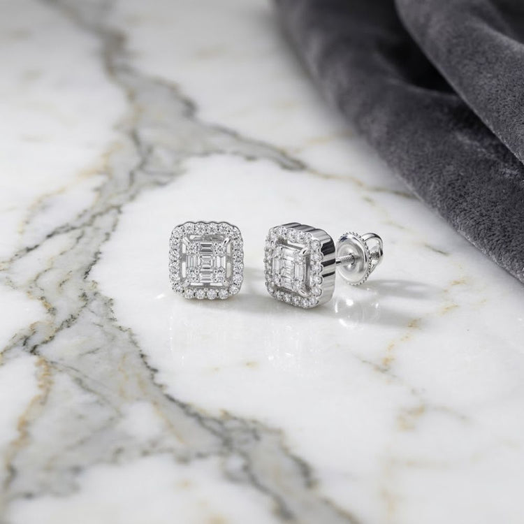 A pair of 1/5 CTW Natural Diamond Baguette Earrings with halo settings rests on a white marble surface beside a folded piece of dark gray velvet fabric.