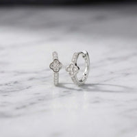 Two 1/10 CTW natural diamond clover hoop earrings, crafted in silver and featuring flower-shaped center designs, are elegantly displayed on a white marble surface.